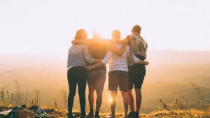 four people with their arms around each others' necks and waists, with their backs to us, at the top of a rise- they are looking at the sunset