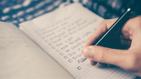 A hand holding a pen, going through a set of checklist items in a small graph paper notebook