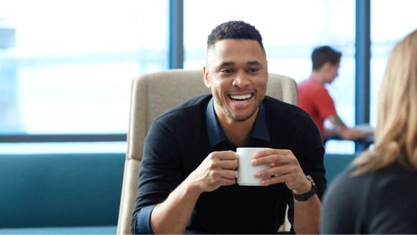 A male teammate drinking coffee, seated, and smiling, facing the camera. We see the back of another teammate with reddish hair and a dark colored shirt.