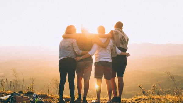 four people with their arms around each others' necks and waists, with their backs to us, at the top of a rise- they are looking at the sunset