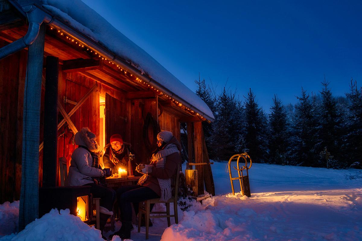People sitting by fire in front of log cabin.