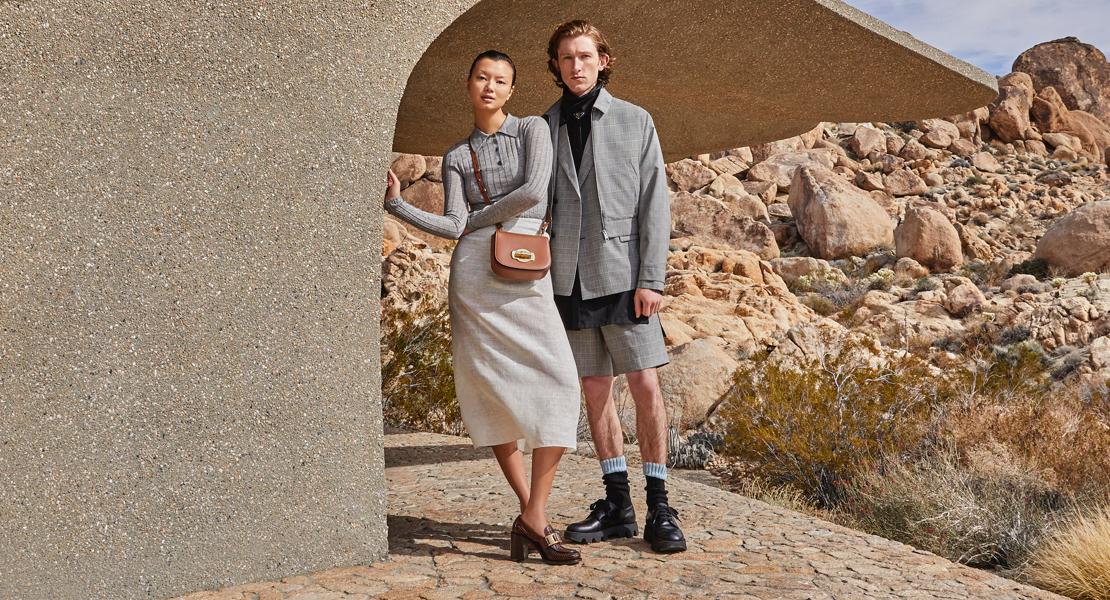 Zwei Models, gekleidet in pastellfarbener und grauer Luxusmode, in einer Wüstenlandschaft.
