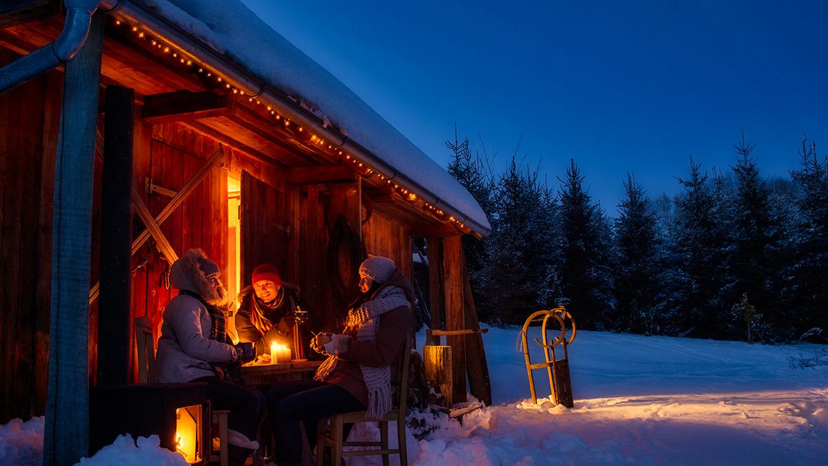 People sitting by fire in front of log cabin.