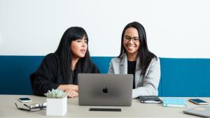 Two women colleagues sitting in front of a Mac laptop: one woman on the left with a black shirt, and another woman on the right with glasses and a gray blazer
