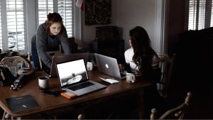 Two coworkers in a darkened office working on their computers, seated around a table. One worker is standing, next to another who is seated at the head of the table.