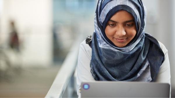 A user on their laptop, facing us - wearing a darker gray and light blue hijab and a lighter colored long sleeve shirt