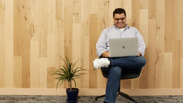 a teammate sitting cross legged in a swivel chair with a small plant at their feet, the teammate is looking at their keyboard and smiling. There is a wood paneled background behind.
